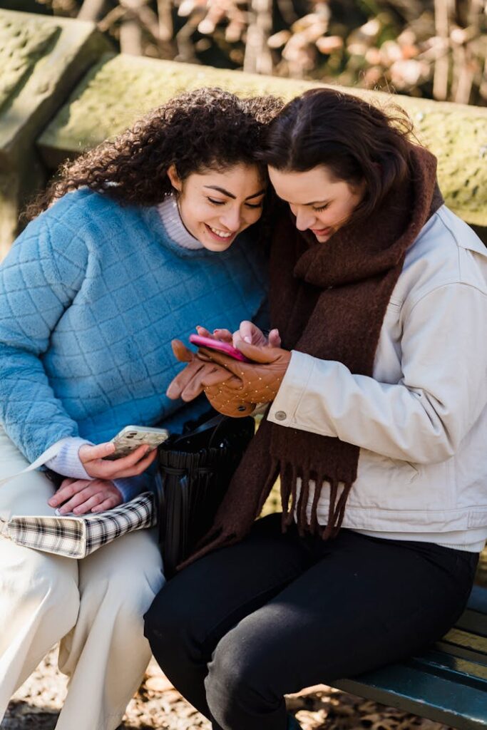 Two women sitting on a park bench, smiling and browsing smartphones together.
